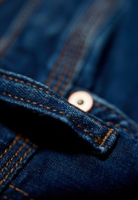 Denim w głębokim niebieskim kolorze z widocznym kontrastowym brązowym przeszyciem. Charakteryzuje się detalem guzika z czarnym środkiem. Gładka tekstura.