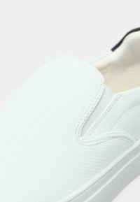 Chaussures blanches à enfiler avec une tige texturée, des inserts élastiques sur les côtés et une semelle en caoutchouc lisse, présentant un design minimaliste et des lignes épurées.