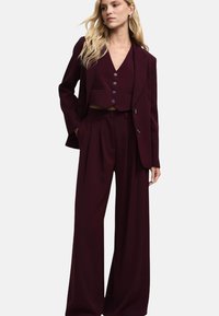 Completo bordeaux composto da gilet corto con bottoni e pantaloni a gamba larga. Il tessuto ha una texture liscia con elementi di design sartoriale.