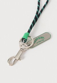 Μπρελόκ με πλεγμένο κορδόνι σε μαύρο και πράσινο χρώμα και ασημένιο μεταλλικό κούμπωμα, με μια ταμπέλα χαραγμένη με τη φράση "CALVIN KLEIN New York City" με πράσινα γράμματα.