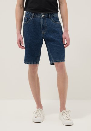 Hombre con pantalones cortos de mezclilla azul oscuro, camiseta negra y zapatillas blancas, de pie contra un fondo liso y claro.