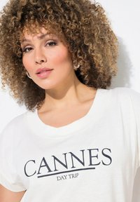 Camiseta blanca con la inscripción "CANNES DAY TRIP" en letras gruesas de color azul marino. Tela suave, corte relajado y escote redondeado.