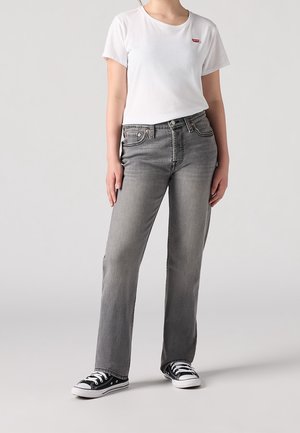 Person iført en hvid Levi's T-shirt, grå jeans med lige ben og sorte lave sneakers, stående mod en ensfarvet baggrund.