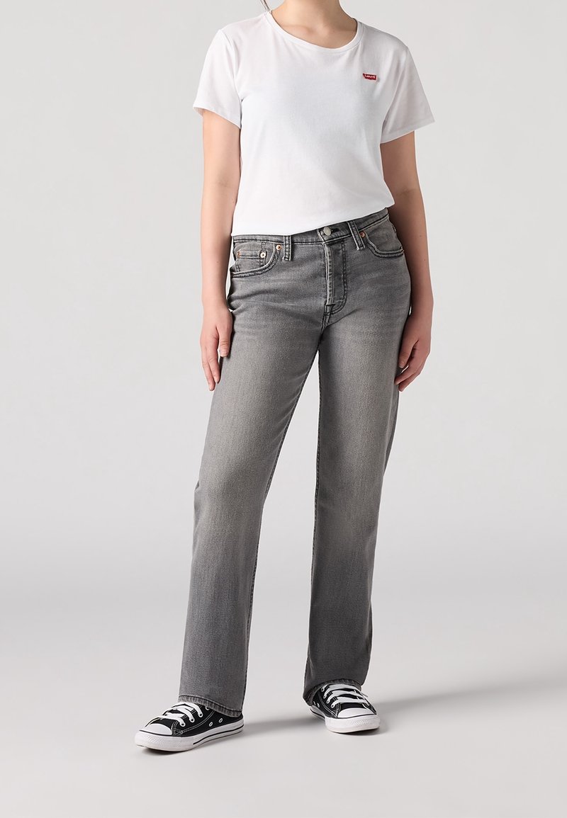 Personne portant un T-shirt blanc Levi's, un jean droit gris et des baskets basses noires, debout devant un fond uni.