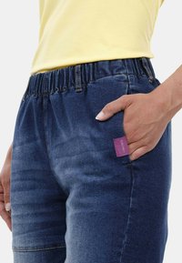 Joggers de mezclilla con cintura elástica, color azul oscuro, y un bolsillo lateral que presenta una etiqueta de marca morada. Tela texturizada con un ligero desgastado.