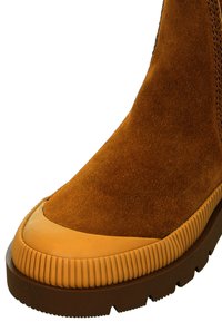 Brown suede ankle boot with rubber sole, featuring a smooth, tan leather toe cap and textured ridged accents along the sole edge.