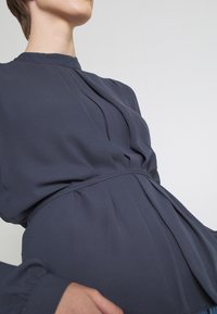 Dunkelgraue Bluse mit hohem Kragen, gefaltetem Vorderteil und tailliertem Schnitt. Weicher Stoff mit langen Ärmeln und gerafften Akzenten.