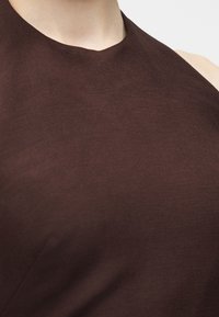 Κοντινό πλάνο καφέ τοπ χωρίς μανίκια με ψηλό, ασύμμετρο λαιμόκοψη σε ένα άτομο μπροστά σε απλό φόντο.