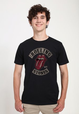 Giovane uomo con capelli ricci che indossa una maglietta nera dei Rolling Stones con il logo della lingua rossa, sorridente su uno sfondo bianco uniforme.