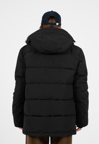 Černá péřová bunda s kapucí, s prošívanými panely a matným povrchem. Zadní strana má volný střih a minimální detaily hardware.