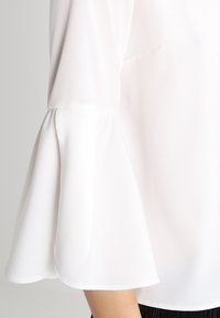 Blusa bianca leggera con maniche a campana. Il materiale appare liscio e leggermente trasparente. Presenta un design rilassato con cuciture minime.