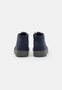 Chaussures bleu foncé montant à la cheville avec des semelles gris tachetées, vues de dos sur un fond blanc uni.