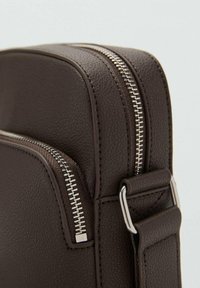 Borsa in pelle marrone con superficie testurizzata, dotata di zip argentate e una tracolla removibile. Forma rettangolare compatta, design minimalista.