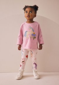 Rosa langärmeliges Shirt mit Einhorn-Stickerei, kombiniert mit weißen Leggings mit bunten Tier- und Blumenmustern. Weiße Turnschuhe.