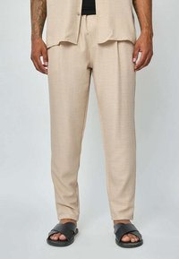 Pantalons beiges légers avec une coupe relaxe, texture subtile et poches latérales, associés à des sandales noires à enfiler.