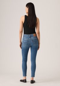 Blaue Skinny-Jeans aus Denim mit hoher Taillenhöhe, ausgestattet mit Gesäßtaschen und einem braunen Leder-Patch. Das Model trägt schwarze Ballerinas.