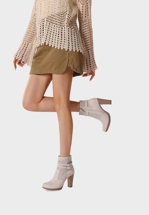 Donna con stivaletti beige traforati con tacco a blocco, una minigonna marrone e un maglione di crochet color crema, in piedi su una gamba sola.