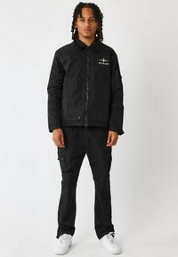 Sæt med sort yderbeklædning bestående af en zip-up jakke med patch lommer og krave, parret med cargo bukser. Begge dele er lavet af tekstureret stof.