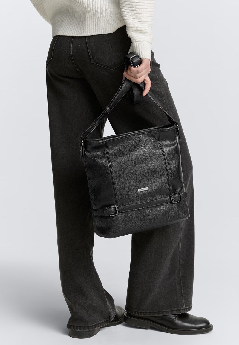 Borsa a tracolla nera in pelle con superficie testurizzata, cintura regolabile e dettagli con doppia fibbia, tenuta da una persona che indossa pantaloni scuri.