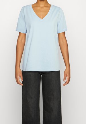 Persona vestida con una camiseta de mangas cortas en V color azul claro y unos pantalones vaqueros rectos negros, de pie frente a un fondo blanco liso.