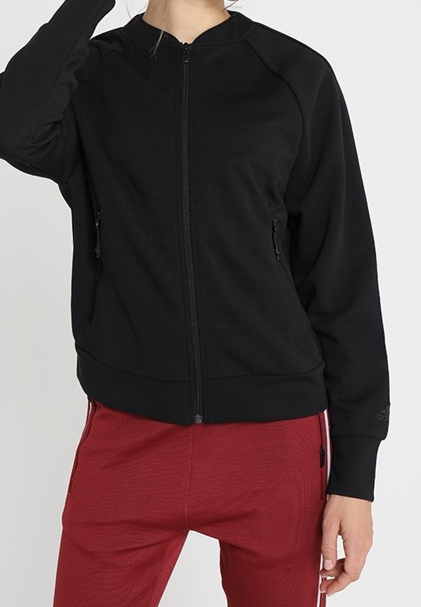 Chaqueta de cremallera negra hecha de tela suave, con cuello redondo, dos bolsillos laterales y puños de canalé. Combinada con pantalones rojos.