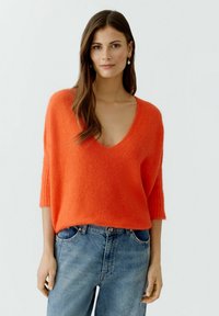 Orangefarbener Wollmischpullover mit tiefem V-Ausschnitt und kurzen, gerippten Ärmeln, kombiniert mit hellblauen Denim-Jeans. Weicher Stoff, legere Passform.