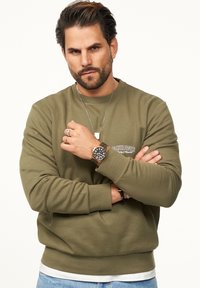 Olive green sweatshirt with ribbed texture and crew neckline, featuring a small logo on the chest. Paired with blue jeans and a watch.