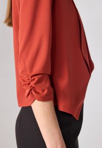 Top de color óxido con mangas de tres cuartos, que presenta detalles de fruncido en los puños y un tejido suave y texturizado. Llevado con pantalones negros.