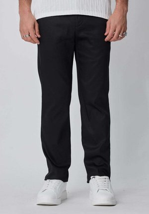 Homme portant un pantalon noir, des baskets blanches et une chemise blanche avec des bagues aux doigts, debout devant un fond clair uni.