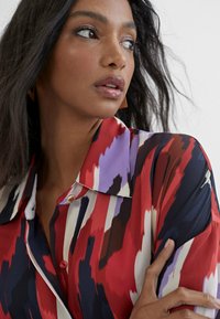 Blusa a motivi rossa, viola e blu navy con colletto, dettagli con bottoni di perla e una consistenza liscia. La mano della modella poggia sul braccio, con orecchini di legno visibili.