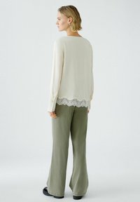 Langärmlige cremefarbene Bluse mit Spitzenbesatz am Saum, kombiniert mit olivgrünen, weiten Hosen und klobigen schwarzen Schuhen. Glattes Material, lässige Passform.