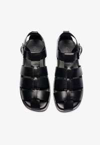 Sandalias de cuero negro con un diseño trenzado, correa ajustable con hebilla y suela plana, con una textura suave y forma minimalista.