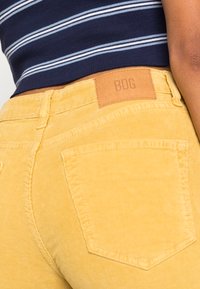 Calças de cotim amarelas com uma superfície texturizada, cinco bolsos e um emblema em pele castanha com "BDG" estampado na parte de trás.