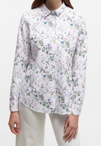 Woman wearing long-sleeve white button-up shirt with pink and blue floral and bird pattern, paired with white pants.
