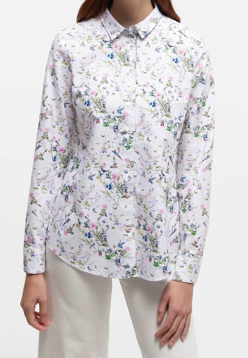 Woman wearing long-sleeve white button-up shirt with pink and blue floral and bird pattern, paired with white pants.