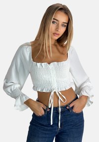 Hvid cropped bluse med smocket front, flæset halsudskæring og lange, elastiske ærmer. Kombination med højttaljede blå jeans.