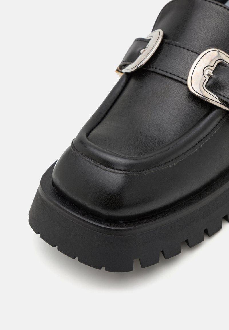 Loafer en cuir noir avec un bout carré, présentant deux boucles en argent et une semelle en caoutchouc épaisse avec une texture striée.