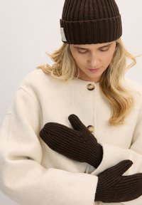 Brązowe rowkowane dziergane rękawiczki noszone z kremowym wełnianym płaszczem z okrągłymi guzikami. Projekt podkreśla fakturę i ciepło.