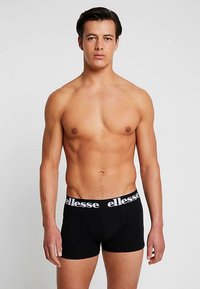 Chiloti boxer negri cu logo alb "ellesse" pe talie, design strâns, material moale și lungime până la mijlocul coapsei. Modelul pozează din față.