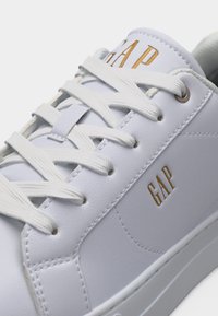 Λευκό δερμάτινο αθλητικό παπούτσι με λευκά κορδόνια και χρυσό λογότυπο "GAP" στη γλώσσα και την πλευρά, εμφανίζοντας λεπτομερείς ραφές και μεταλλικά μυκτήρια.