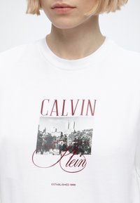 Κοντινό πλάνο ενός ατόμου που φοράει λευκό T-shirt με ασπρόμαυρο τοπίο πόλης και κόκκινο κείμενο που γράφει Calvin Klein Established 1968.