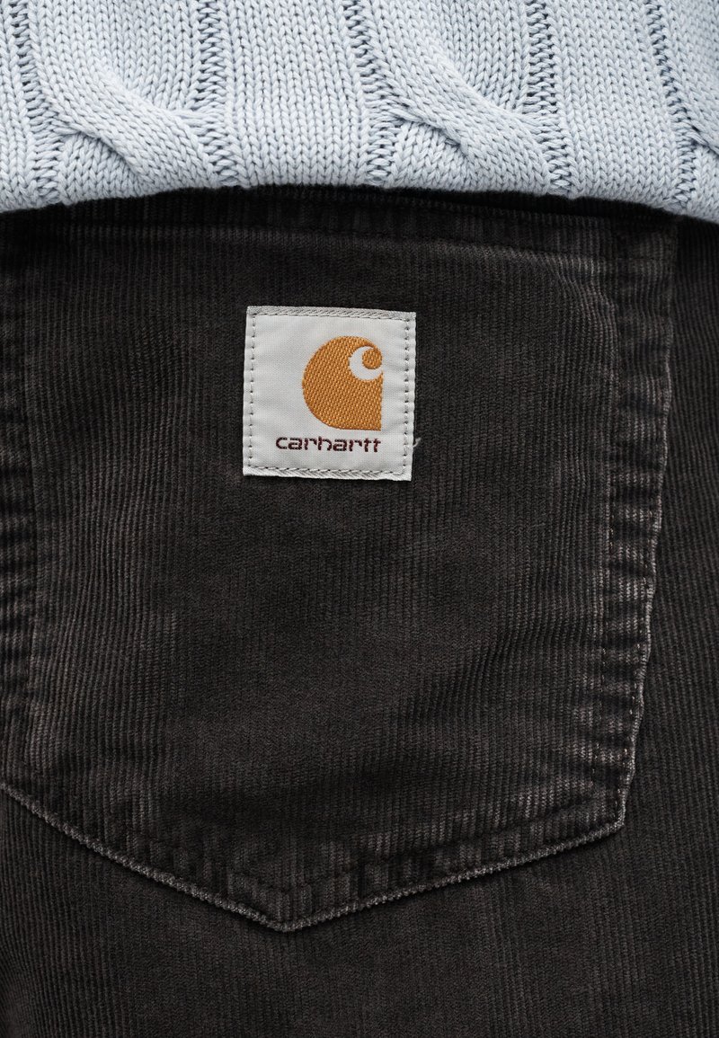 Poche en velours côtelé foncé avec une étiquette Carhartt blanche cousue dessus, sous un pull en maille torsadée bleu clair.