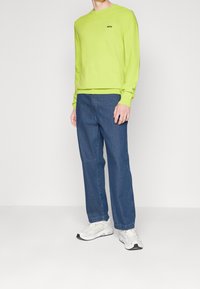 Man som bär en limegrön långärmad tröja med "BOSS"-logotyp, blå jeans och vita sneakers, stående mot en enkel vit bakgrund.