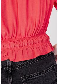 Blusa corallo brillante con orlo elastico arricciato, tessuto leggero e superficie liscia, abbinata a jeans in denim neri con tasca posteriore.