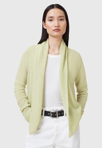 Cardigan aperto di un verde pallido con una texture morbida, maniche lunghe e due tasche frontali, abbinato a una maglia bianca e una cintura nera.