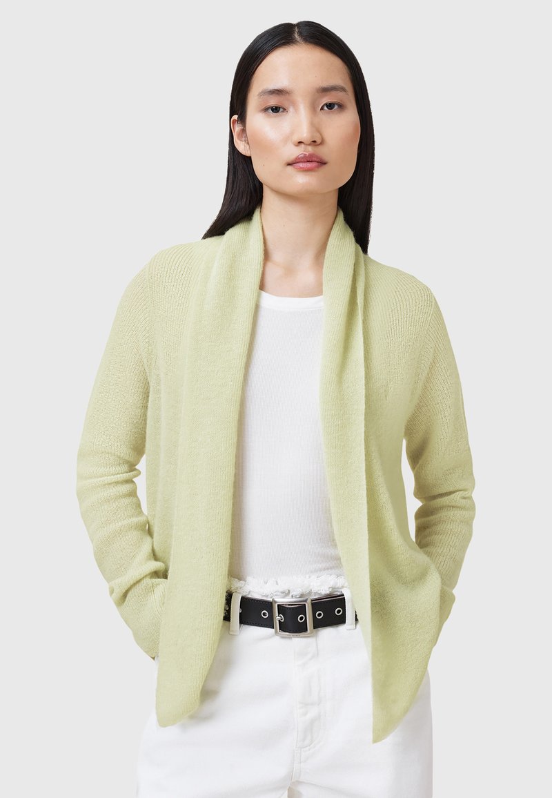Cardigan aperto di un verde pallido con una texture morbida, maniche lunghe e due tasche frontali, abbinato a una maglia bianca e una cintura nera.