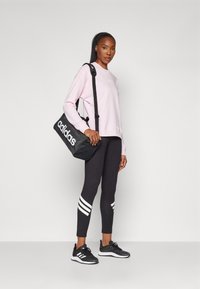 Sudadera rosa con logo blanco, leggings negros con rayas blancas, zapatillas deportivas negras y un bolso de hombro negro con marca blanca.