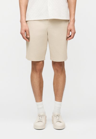 Homme portant un short beige jusqu'aux genoux, des chaussettes blanches et des baskets beiges, sur un fond blanc uni.