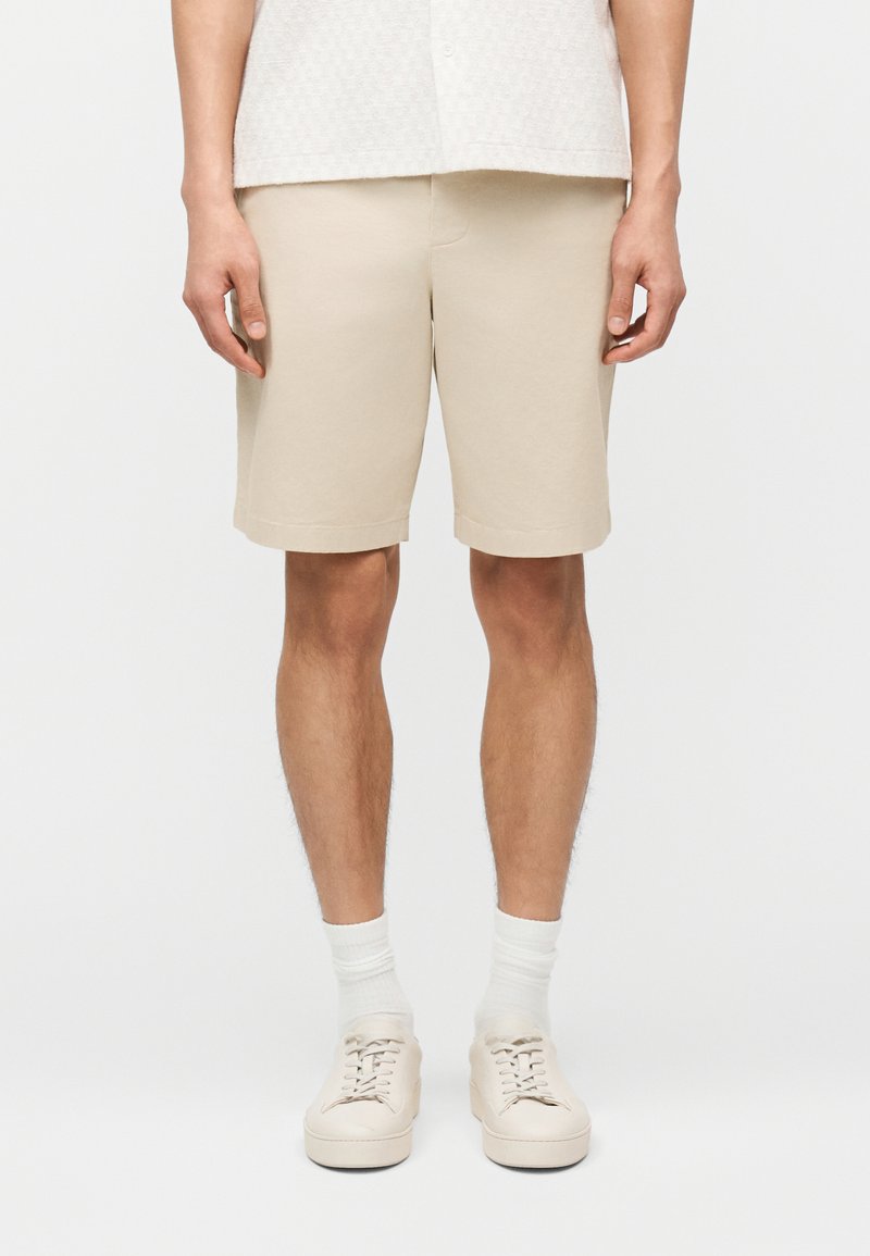 Άντρας που φοράει μπεζ σορτς μέχρι το γόνατο, λευκές κάλτσες και μπεζ αθλητικά παπούτσια με λευκό φόντο.