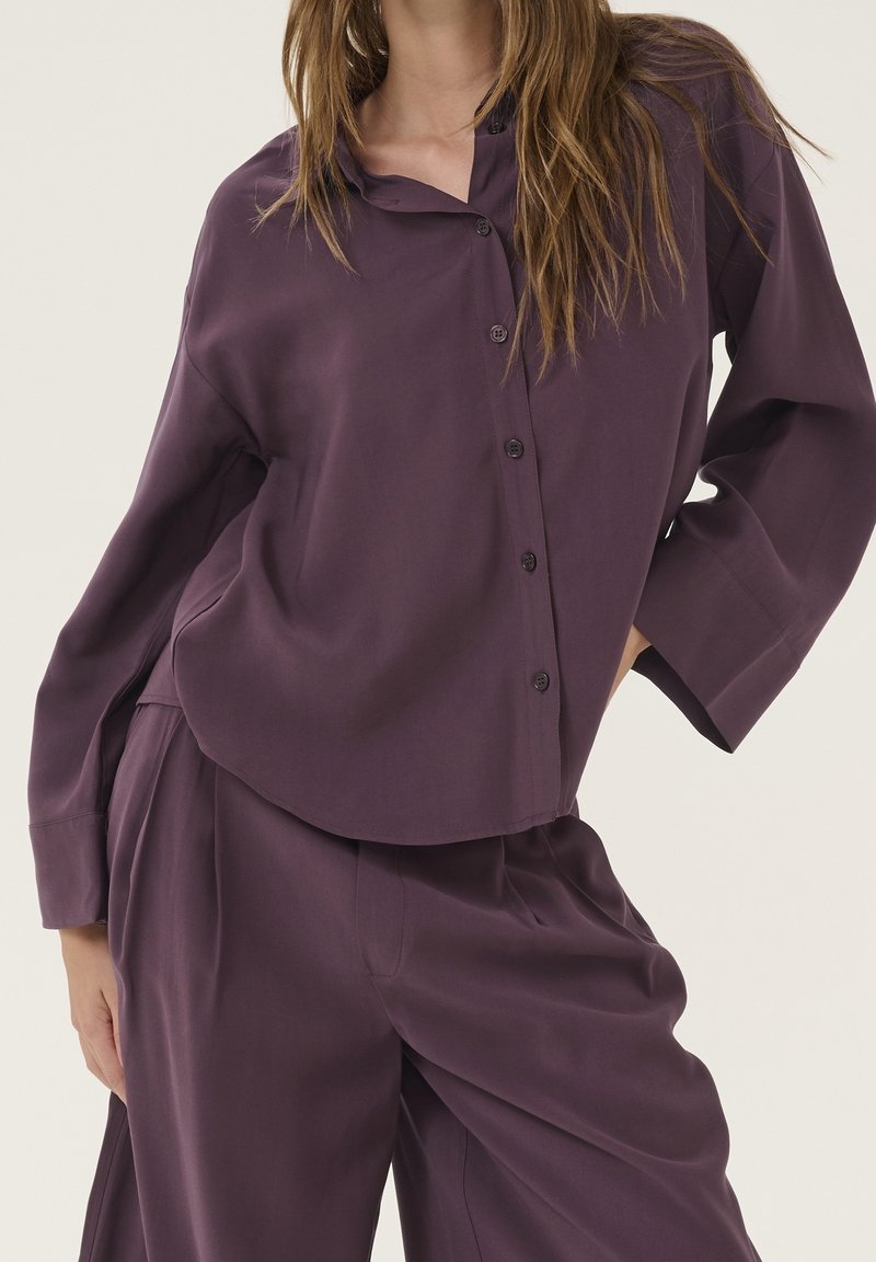 Blusa viola con vestibilità rilassata, maniche lunghe, chiusura con bottoni frontale e orlo arrotondato. Abbinata a pantaloni larghi coordinati. Tessuto morbido.